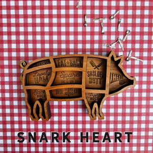 May include: Wooden pig-shaped serving tray with sections labeled for different cuts of meat, including "Sirloin", "Lovely Ham", "Spare Ribs", "Fat + Back", "Shoulder Butt", "Head", "Back Hock", and "Front Hock". The tray is on a red and white checkered background. The text "SNARK HEART" is printed below the tray.