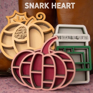 May include: Three wooden layered ornaments in the shape of pumpkins. The first pumpkin is natural wood with the words "pumpkin spice everything nice" inside. The second pumpkin is a deep red with a pink interior. The third ornament is a white coffee cup with green interior and the words "It's basically free #GirlMath" on the side.