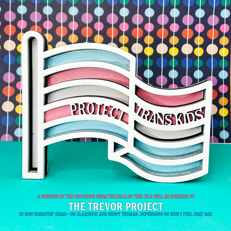 May include: A wooden cutout of a transgender pride flag with the words "Protect Trans Kids" on it. The flag is painted in the colors of the transgender pride flag: light blue, pink, white, light blue, pink. The flag is on a turquoise background.