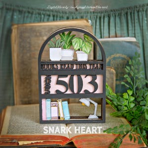May include: A black wooden bookshelf with the text "BOOKS READ THIS YEAR" and the number "503". The shelf features potted plants, books, and a lamp. The text "SNARK HEART" is at the bottom.