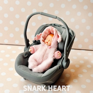 Op de afbeelding: Een miniatuur babypopje in een roze onesie met capuchon en wanten, genesteld in een grijze autostoel. De capuchon heeft een wit stippenpatroon. De tekst "SNARK HEART" is zichtbaar onderaan de afbeelding.