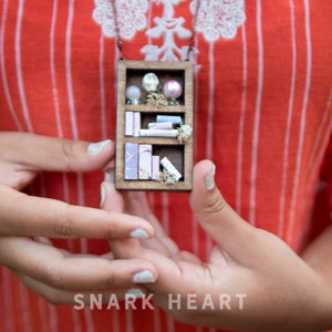 Puede incluir: Un collar de madera con una mini biblioteca en miniatura con un esquema de color rosa y morado. La biblioteca está llena de pequeños libros, una calavera y una perla.