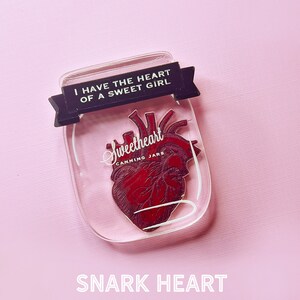 May include: A clear glass jar with a red anatomical heart inside. The jar has a black ribbon with the text "I have the heart of a sweet girl" on it. The jar also has the text "Sweetheart Canning Jars" on it.