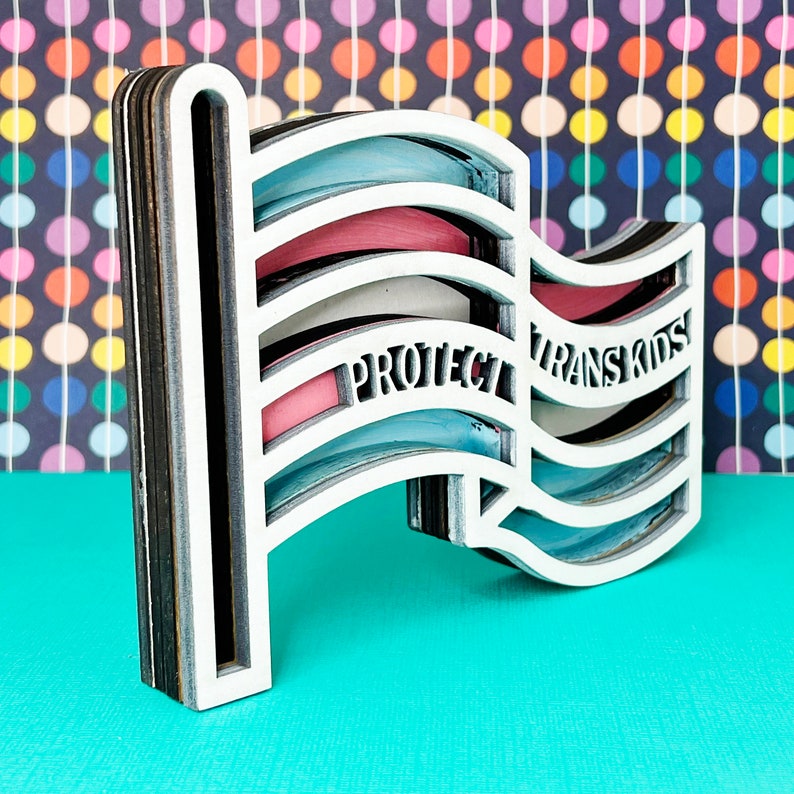 May include: A white wooden cutout of a flag with the colors of the transgender pride flag. The flag is waving and has the words "Protect Trans Kids" written on it.