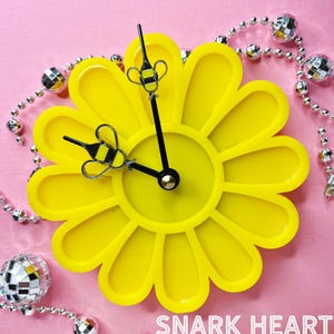 May include: A yellow daisy-shaped clock with black hands and bee accents. The clock is on a pink background with silver beads.