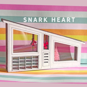 May include: A white miniature house with a slanted roof, featuring large windows and a small pink Christmas tree inside. The text "SNARK HEART" is displayed above the house against a striped background.