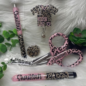 May include: A set of personalized medical accessories in a pink and black leopard print. The set includes a pen, a badge holder, a pair of scissors, and a small round button. All items are decorated with glitter and the name "Shaileah" is written on each item. The badge holder has the text "Scrub Life" on it.