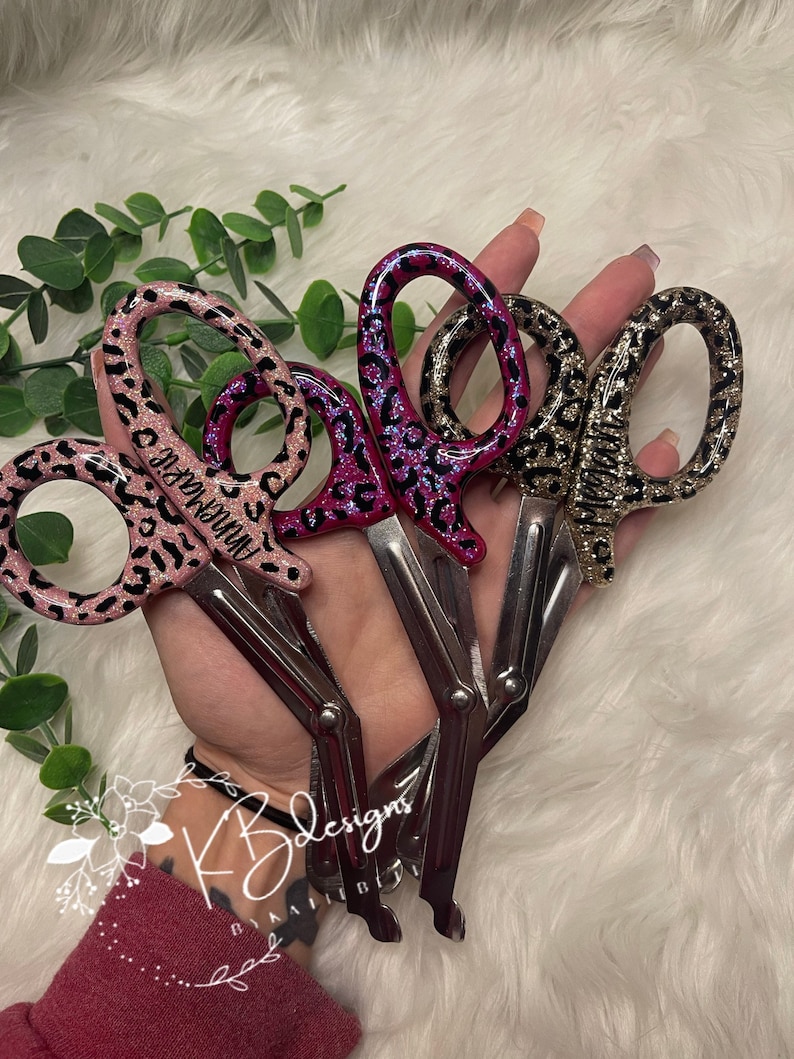 Op de afbeelding: Drie paar chirurgische scharen met luipaardprint handvatten. De handvatten zijn roze, paars en goud. De scharen zijn gesloten.