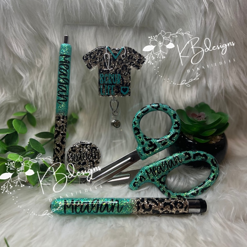 May include: A set of teal and black leopard print accessories for nurses. The set includes a pen, a badge reel, a pair of scissors, and a retractable pen. The accessories are personalized with the name "Meagan".