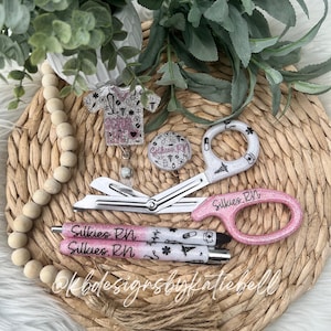 May include: Pink and white glitter nurse themed retractable badge reel with a pair of pink scissors with the text "Silkies RN" and two pink pens with the text "Silkies RN".