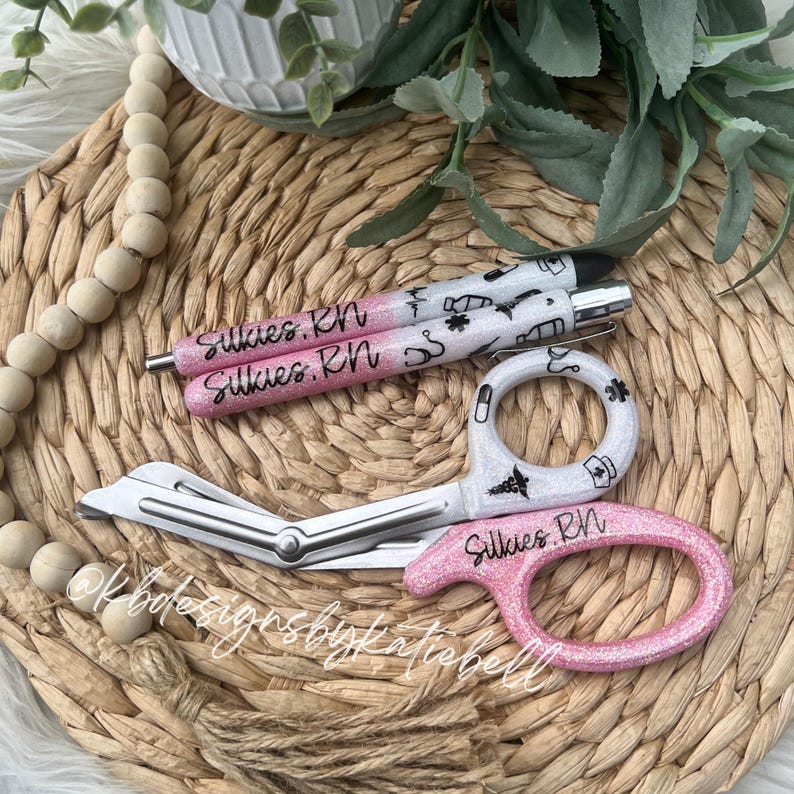 May include: Two pink and white glitter pens with the text "Silkies. RN" and a pair of pink and white glitter medical scissors with the text "Silkies. RN".