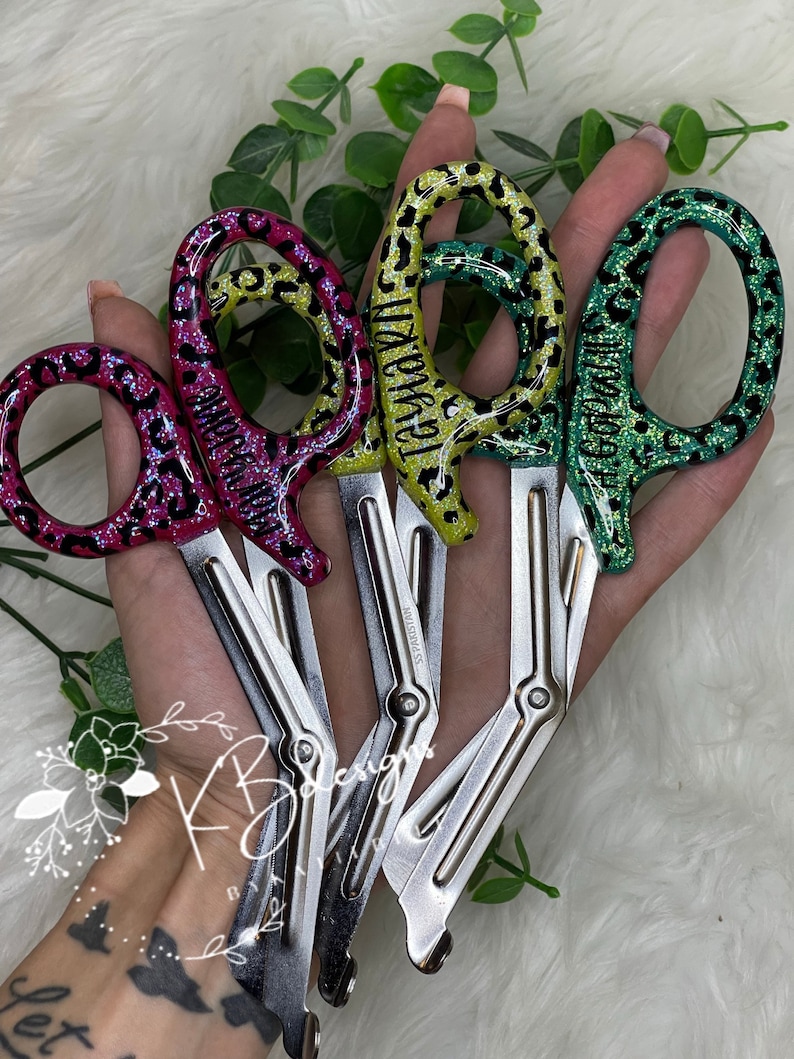 Op de afbeelding: Vier paar zilveren medische scharen met zwarte en groene luipaardprint handvatten. De handvatten zijn versierd met glitter en hebben de woorden "Teagan", "Morgan", "Taylor" en "Madison" erop geschreven.