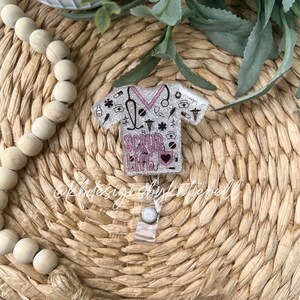 May include: A silver glitter scrub top with pink accents and the text "Scrub Life" is attached to a clear badge holder. The scrub top is decorated with medical symbols and a heart.