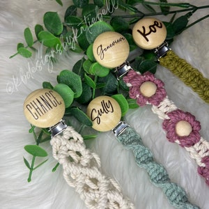 May include: Four wooden baby teethers with different names engraved on them. Each teether is attached to a crocheted pacifier clip in a different color: cream, green, pink, and brown.