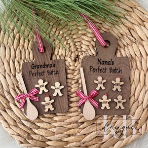 May include: Two wooden cutting board ornaments with the text "Grandma's Perfect Batch" and "Nana's Perfect Batch". Each ornament has gingerbread people and a wooden spoon with a red and white ribbon. The ornaments are hanging from a red and white checkered ribbon.