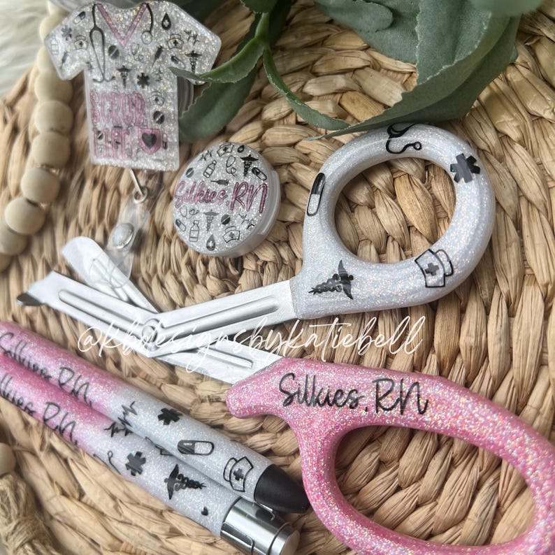 May include: A set of medical themed accessories including a pair of silver glitter scissors, a pink glitter pen with the text "Silkies RN", a silver glitter badge reel with a pink glitter charm, and a silver glitter button with the text "Silkies RN".