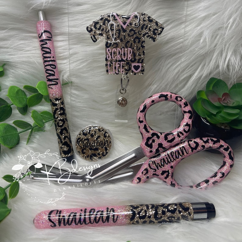 May include: A set of personalized medical accessories in a pink and black leopard print. The set includes a pen, a badge holder, a pair of scissors, and a small round button. All items are decorated with glitter and the name "Shaileah" is written on each item. The badge holder has the text "Scrub Life" on it.