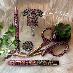 May include: A set of pink and black leopard print medical accessories, including a pen, scissors, badge reel, and a button. The badge reel features a black and gold glitter shirt with the text "Scrub Life".