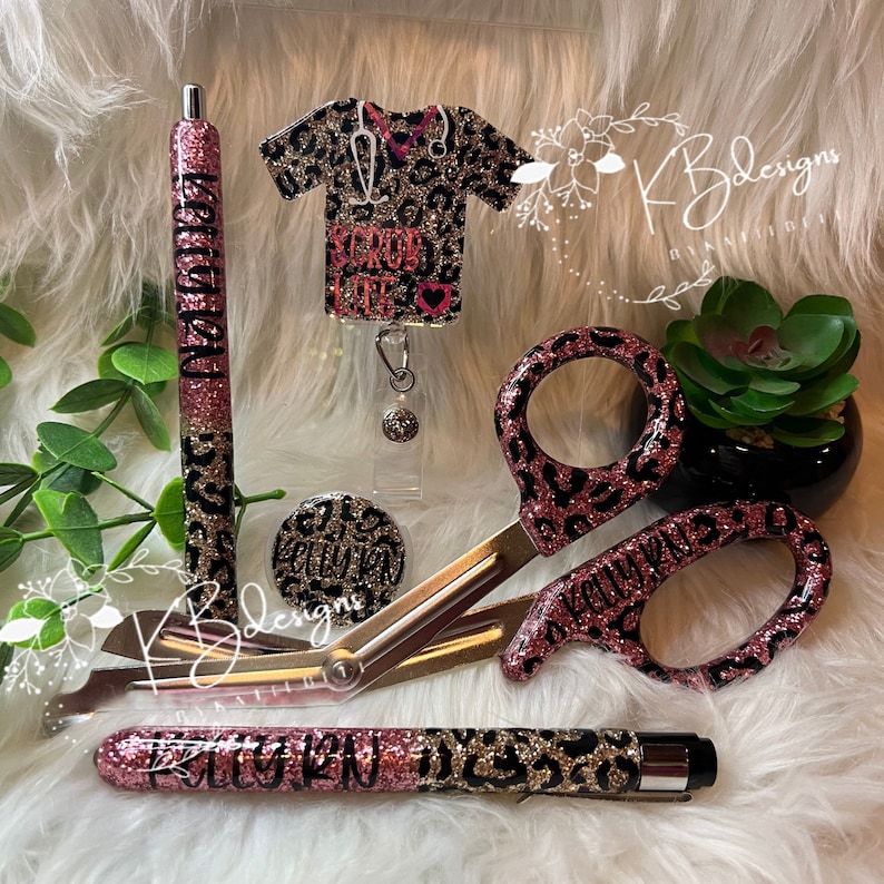 May include: A set of pink and black leopard print medical accessories, including a pen, scissors, badge reel, and a button. The badge reel features a black and gold glitter shirt with the text "Scrub Life".