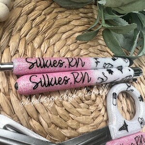 May include: Two pink and white glitter pens with black lettering that says "Sulkie's RN". The pens have a white background with black medical symbols.