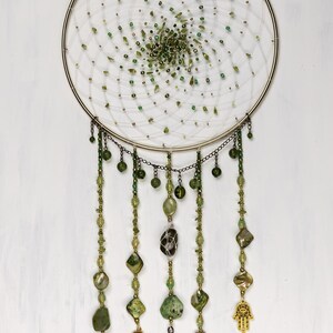 May include: A gold dreamcatcher with a white net and green beads. The dreamcatcher has five strands of beads hanging from it, each with a different green stone or charm. The charms include a green stone, a green stone with a white design, a green stone with a gold design, a green stone with a gold design, and a gold hamsa hand.