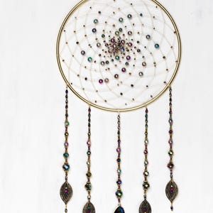 May include: A gold dreamcatcher with a white web and iridescent beads. The dreamcatcher has five strands of beads with iridescent beads and a crystal pendant at the end of each strand.