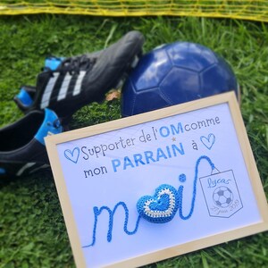May include: A framed sign with a blue crocheted heart and the text "Supporter de l'OM comme mon PARRAIN &agrave;" and "Lucas 27 juin 2019".