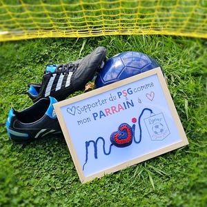 May include: A framed sign with a blue and red crocheted heart and the text "Supporter du PSG comme mon PARRAIN &agrave; moi" with a soccer ball and a pair of soccer cleats in the background.