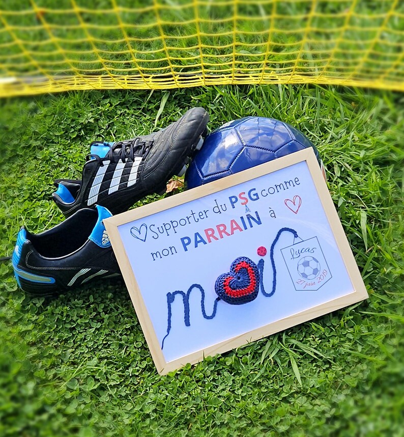 May include: A framed sign with a blue and red crocheted heart and the text "Supporter du PSG comme mon PARRAIN &agrave; moi" with a soccer ball and a pair of soccer cleats in the background.