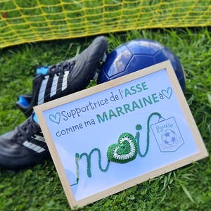 May include: A framed white sign with green yarn lettering that says "Supportrice de l'ASSE comme ma MARRAINE a" with a green yarn heart and a small white sign that says "Louise Juillet 2021". The sign is sitting on green grass with a soccer ball and two black and white soccer cleats.