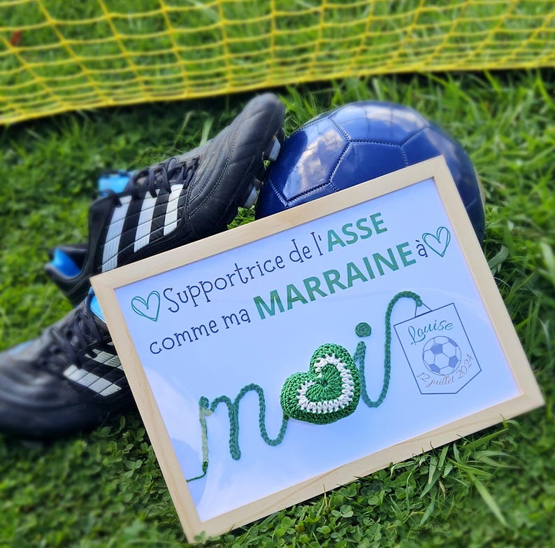 May include: A framed white sign with green yarn lettering that says "Supportrice de l'ASSE comme ma MARRAINE a" with a green yarn heart and a small white sign that says "Louise Juillet 2021". The sign is sitting on green grass with a soccer ball and two black and white soccer cleats.