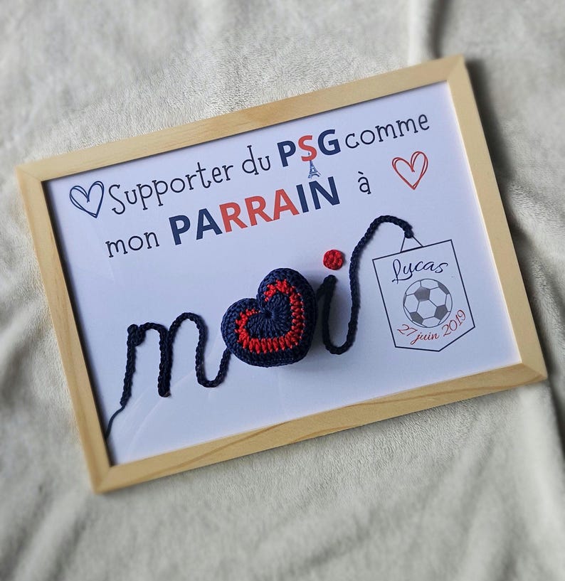 May include: A framed white board with the text "Supporter du PSG comme mon PARRAIN &agrave;" and a blue and red crocheted heart with the word "moi" written in blue yarn. The board also has a small white card with the text "Lucas 27 juin 2019" and a soccer ball graphic.