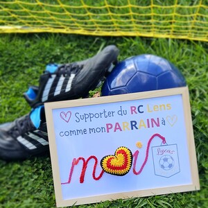 May include: A framed sign with a crocheted heart that says "Supporter du RC Lens comme mon PARRAIN &agrave; moi" with a soccer ball in a pocket that says "Lucas".