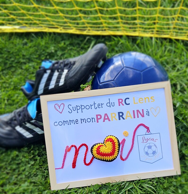 May include: A framed sign with a crocheted heart that says "Supporter du RC Lens comme mon PARRAIN &agrave; moi" with a soccer ball in a pocket that says "Lucas".