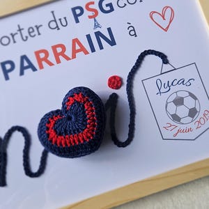 May include: A framed white board with the text "Parrain du PSG comme &agrave;" and a red heart. A blue crocheted heart with a red border is attached to a blue string. A small red crocheted circle is also attached to the string. A smaller framed white board with the text "Lucas 27 juin 2019" and a soccer ball is in the bottom right corner.