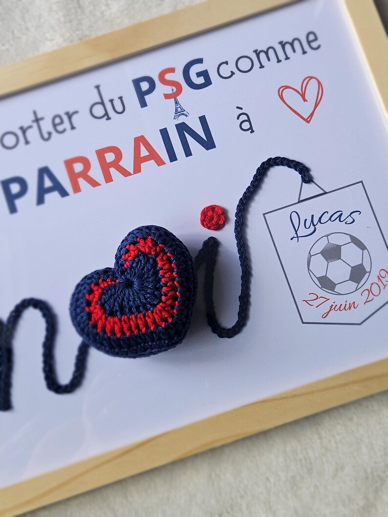 May include: A framed white board with the text "Parrain du PSG comme &agrave;" and a red heart. A blue crocheted heart with a red border is attached to a blue string. A small red crocheted circle is also attached to the string. A smaller framed white board with the text "Lucas 27 juin 2019" and a soccer ball is in the bottom right corner.