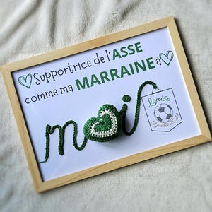 May include: A framed white board with the text "Supportrice de l'ASSE comme ma MARRAINE &agrave;" and a green crocheted heart with the word "moi" spelled out in green yarn. A small white square with the text "Louise 12 juillet 2024" and a soccer ball is in the bottom right corner.