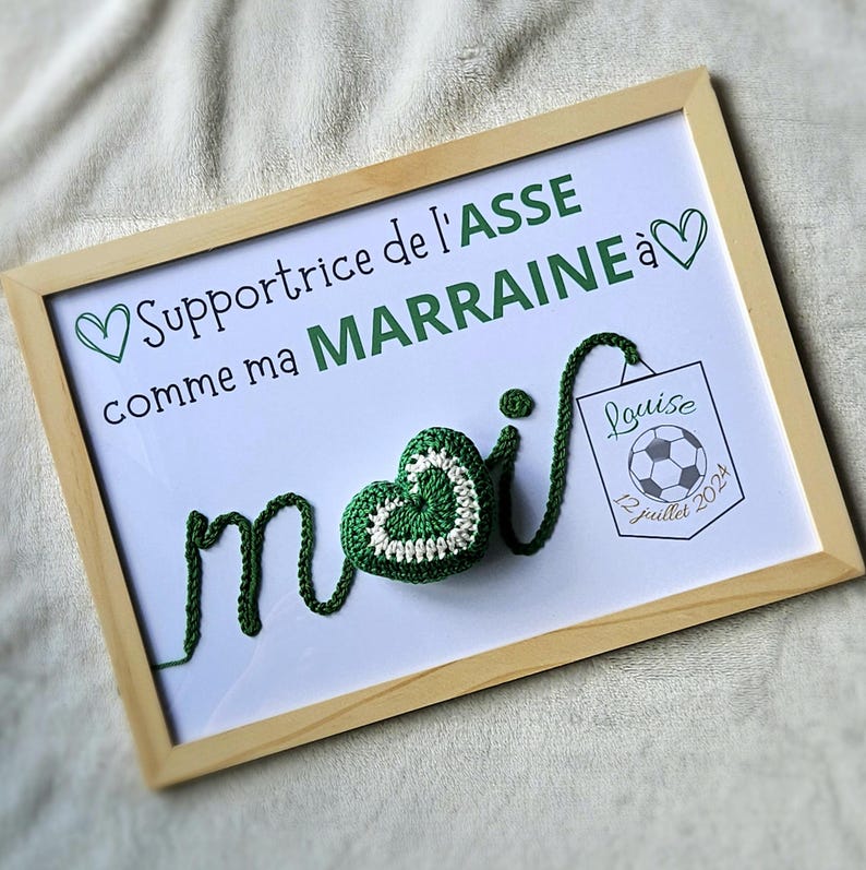 May include: A framed white board with the text "Supportrice de l'ASSE comme ma MARRAINE &agrave;" and a green crocheted heart with the word "moi" spelled out in green yarn. A small white square with the text "Louise 12 juillet 2024" and a soccer ball is in the bottom right corner.