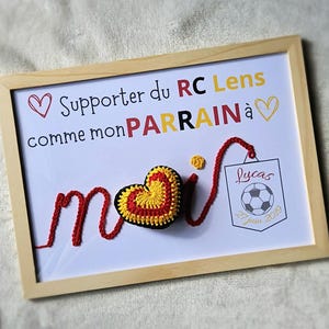 May include: A framed white board with red yarn spelling out "moi" with a red and yellow crocheted heart in the middle. The board has the text "Supporter du RC Lens comme mon PARRAIN &agrave;" with a heart at the beginning and end of the text. A small white tag with the text "Lucas 27 juin 2019" and a black and white soccer ball is attached to the yarn.