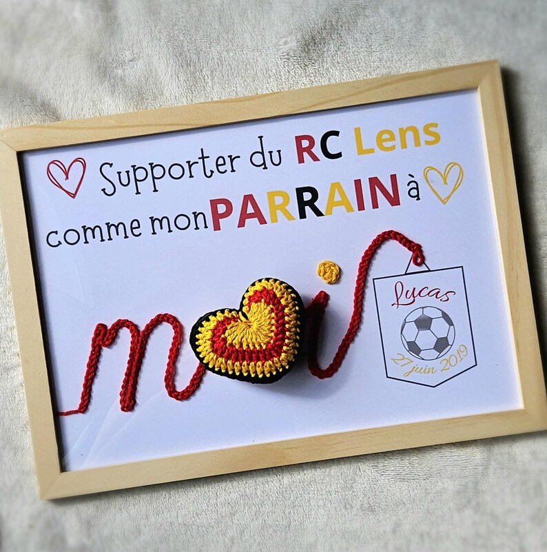 May include: A framed white board with red yarn spelling out "moi" with a red and yellow crocheted heart in the middle. The board has the text "Supporter du RC Lens comme mon PARRAIN &agrave;" with a heart at the beginning and end of the text. A small white tag with the text "Lucas 27 juin 2019" and a black and white soccer ball is attached to the yarn.