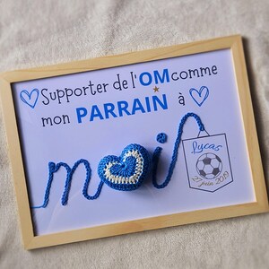 May include: A framed sign with a blue crocheted heart and the text "Supporter de l'OM comme mon PARRAIN &agrave;" and a blue crocheted heart. The sign also has a small tag with the text "Lucas 27 juin 2019" and a soccer ball.