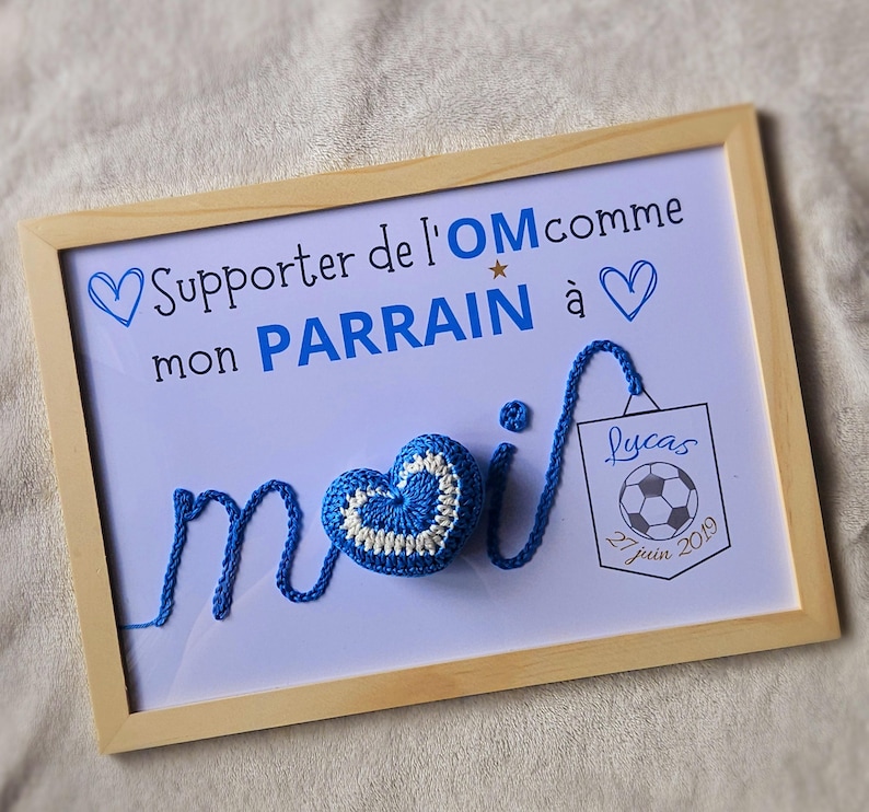 May include: A framed sign with a blue crocheted heart and the text "Supporter de l'OM comme mon PARRAIN &agrave;" and a blue crocheted heart. The sign also has a small tag with the text "Lucas 27 juin 2019" and a soccer ball.