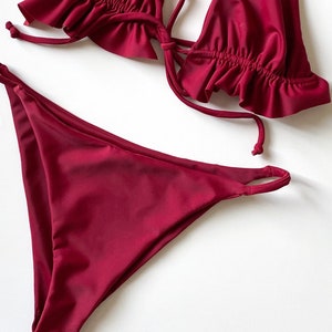 May include: A burgundy two-piece swimsuit. The top has frilled trim and tie straps. The bottoms are a classic bikini style. The set is made from a smooth, shiny fabric.