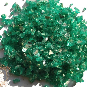 May include: A pile of emerald green crushed glass pieces with reflective surfaces. The irregular shapes and sizes of the glass create a sparkling effect. The image is well-lit, highlighting the color and texture of the glass.