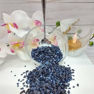 May include: A glass bowl filled with dark blue iridescent glass chips, spilling out onto a white surface. A silver spoon is in the bowl.