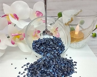 May include: A glass bowl filled with dark blue iridescent glass chips, spilling out onto a white surface. A silver spoon is in the bowl.