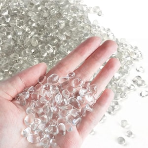 Peut inclure: Une main tenant un tas de galets de verre transparents et lisses. Les galets sont de tailles et de formes variées et sont éparpillés sur un fond blanc.