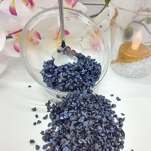May include: A glass bowl filled with dark blue iridescent glass gravel, with a silver spoon with a flower design resting on top of the gravel. The bowl is tipped over, spilling the gravel onto a white surface.