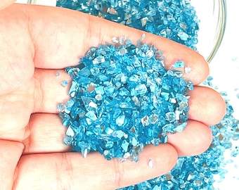 May include: A hand holding a pile of small, iridescent turquoise blue glass pieces. The glass pieces are scattered on a white surface.