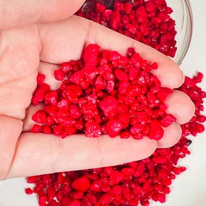 May include: A handful of small, bright red, irregular shaped, decorative glass stones. The stones are scattered on a white surface.